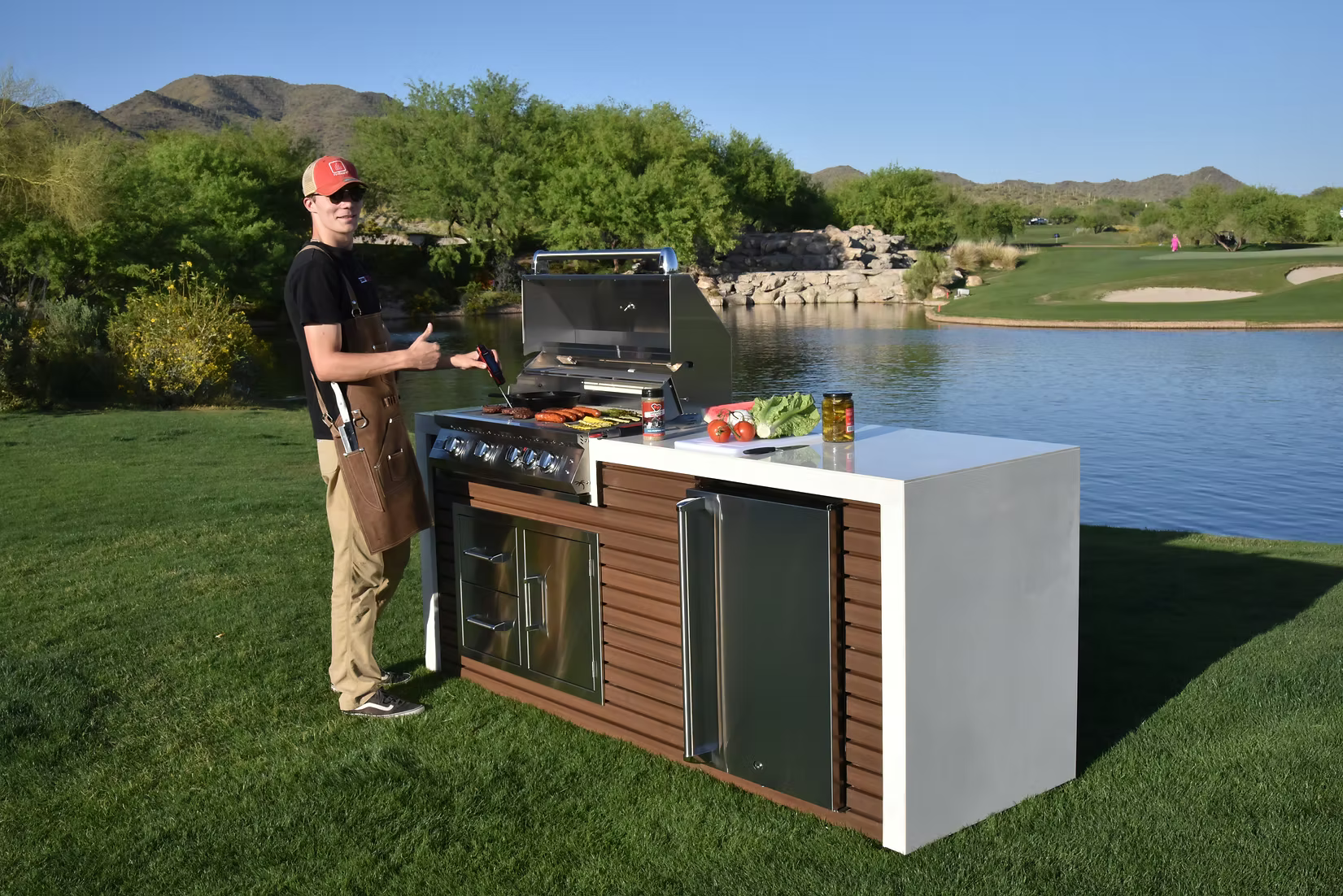 A person in a cap and apron grills food on a Kokomo Grills Professional Shiplap Built-In BBQ Island Outdoor Kitchen with Waterfall Edge by a lake, surrounded by trees, mountains, and a golf course. Ingredients are on the counter.