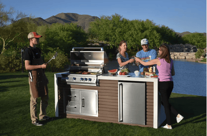Four friends gather by a pond with mountains behind, enjoying drinks and food around a Kokomo Grills Professional Shiplap Built-In BBQ Island Outdoor Kitchen with Waterfall Edge as someone grills at the outdoor kitchen counter.