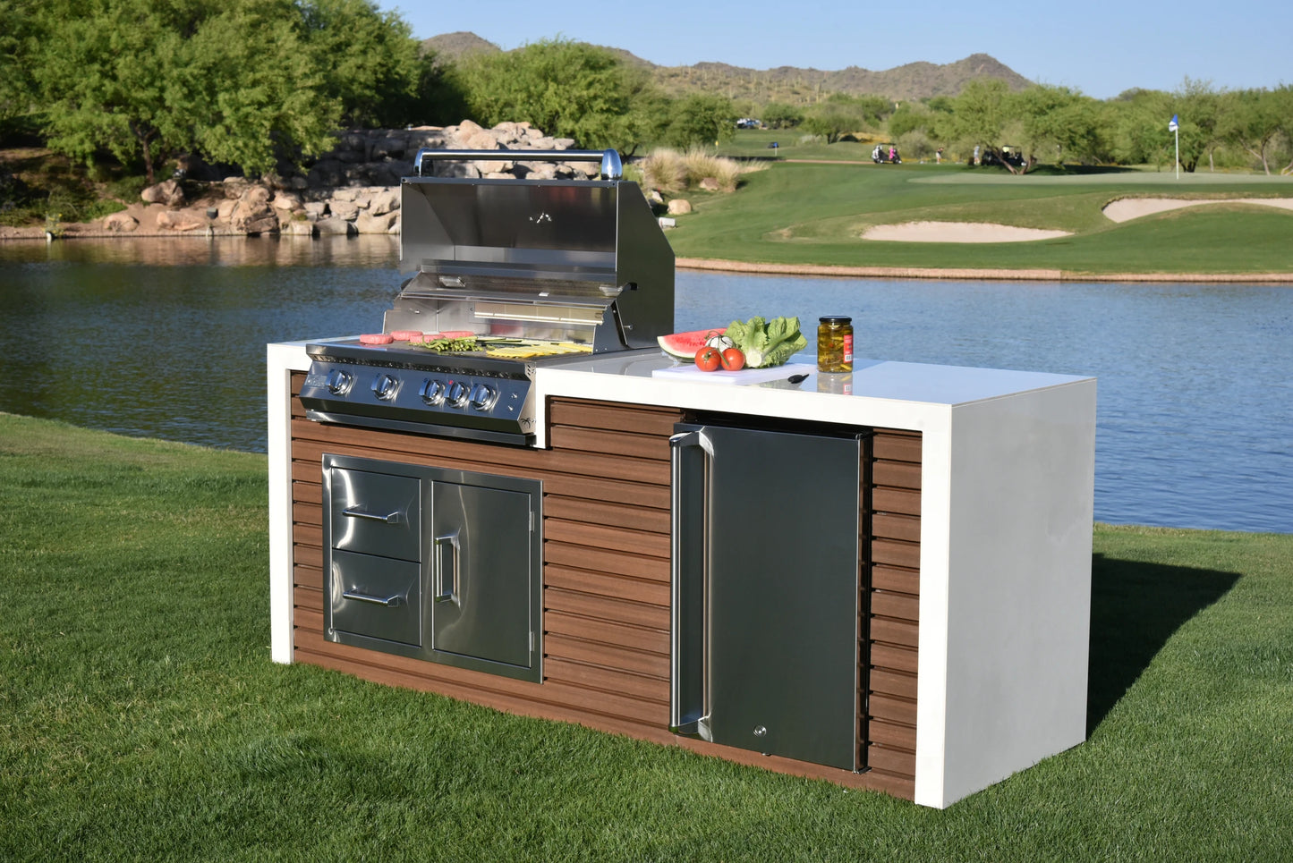 A Kokomo Grills Professional Shiplap Built-In BBQ Island Outdoor Kitchen with Waterfall Edge sits on grass by a lake, with mountains and a golf course behind it. Fresh vegetables and a drink are on the countertop.