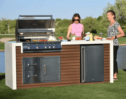Two women prepare food at a Kokomo Grills Professional Shiplap Built-In BBQ Island Outdoor Kitchen with Waterfall Edge, surrounded by vegetables, condiments, and drinks, with a scenic lawn and lake in the background.