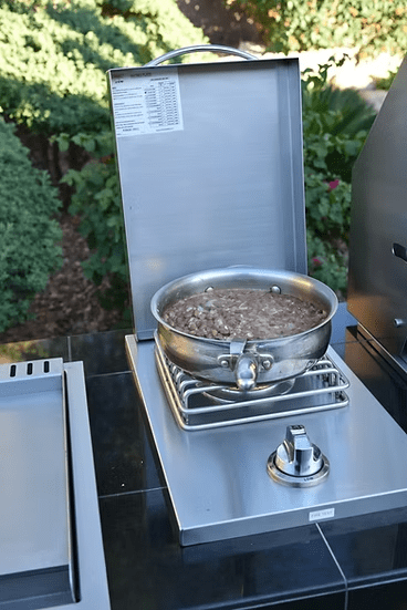 A Kokomo Grills Single Side Burner by Kokomo Grills, crafted from 304 stainless steel, is cooking food in a metal pot outdoors on the patio kitchen, with green plants and bushes in the background.