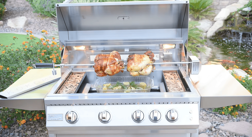 A garden-set stainless steel grill roasts two chickens on a rotisserie over veggies, enhanced by the Kokomo Grills Smoker Chip Box Insert (KO-BAK-SMKBX) for rich flavor, with flowers and rocks nearby.