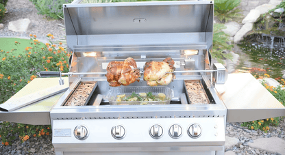 A garden-set stainless steel grill roasts two chickens on a rotisserie over veggies, enhanced by the Kokomo Grills Smoker Chip Box Insert (KO-BAK-SMKBX) for rich flavor, with flowers and rocks nearby.