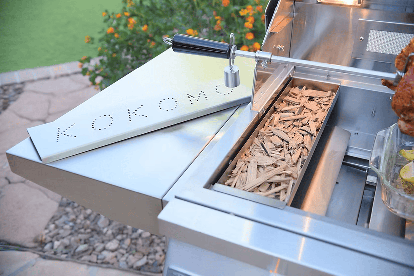 A stainless steel grill showcases the Kokomo Grills Smoker Chip Box Insert (KO-BAK-SMKBX) by Kokomo Grills, a premium BBQ accessory with a hinged “KOKOMO” cover, placed on a stone patio by green landscaping and orange flowers.