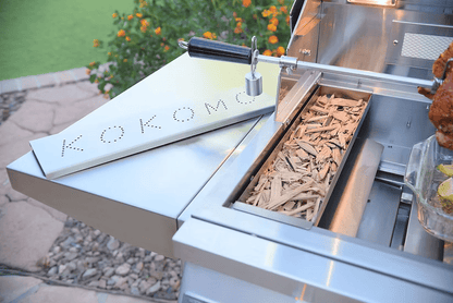 A stainless steel grill showcases the Kokomo Grills Smoker Chip Box Insert (KO-BAK-SMKBX) by Kokomo Grills, a premium BBQ accessory with a hinged “KOKOMO” cover, placed on a stone patio by green landscaping and orange flowers.