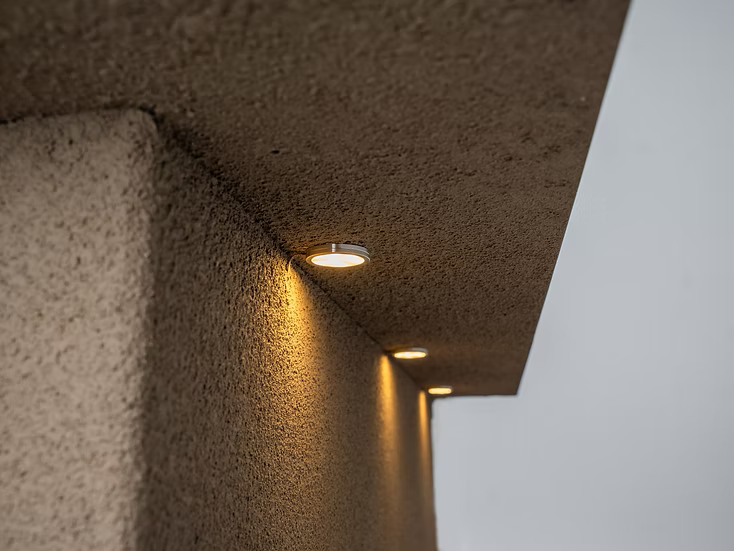 A close-up shows a textured ceiling overhang with recessed round lights casting a warm glow above the Kokomo Grills St. John 7-Foot and 6-Inch Kitchen Island, with a gray sky in the background.