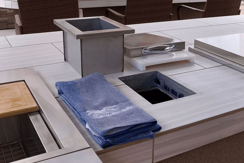 A counter featuring a metal container, two stacked blue towels, a covered food tray, and a Kokomo Grills Trash Chute and Cutting Board KO-TRSCH by Kokomo Grills in the surface, likely for waste or storage, in an outdoor or commercial kitchen setting.