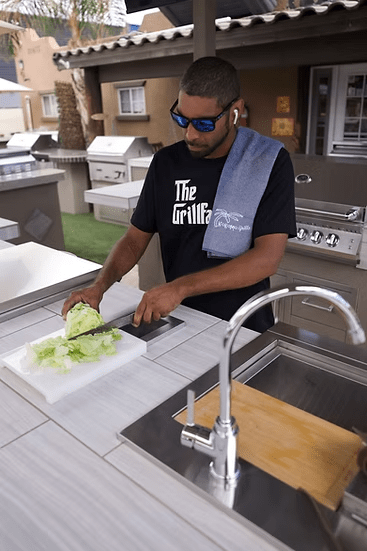 A man in sunglasses and earphones slices lettuce on the Kokomo Grills Trash Chute and Cutting Board KO-TRSCH in an outdoor kitchen, with a towel over his shoulder. He stands beside a sink and grill from Kokomo Grills.