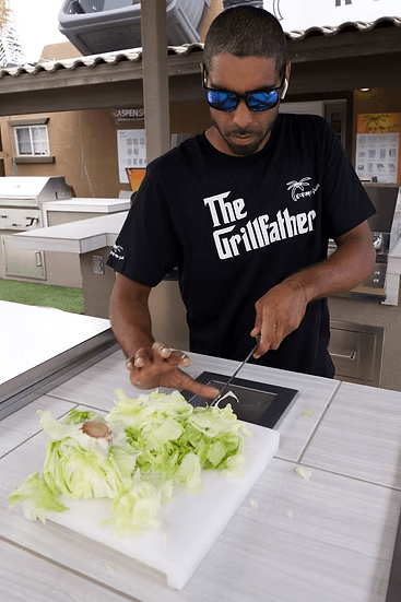 A man in sunglasses and a black The Grillfather t-shirt slices lettuce on a cutting board beside a Kokomo Grills Trash Chute and Cutting Board KO-TRSCH in an outdoor kitchen area.