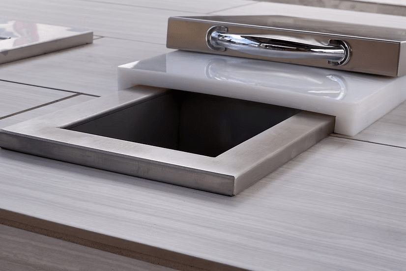 The Kokomo Grills Trash Chute and Cutting Board KO-TRSCH by Kokomo Grills is built into a tiled countertop, featuring a modern square design with a shiny lid—ideal for any outdoor kitchen.