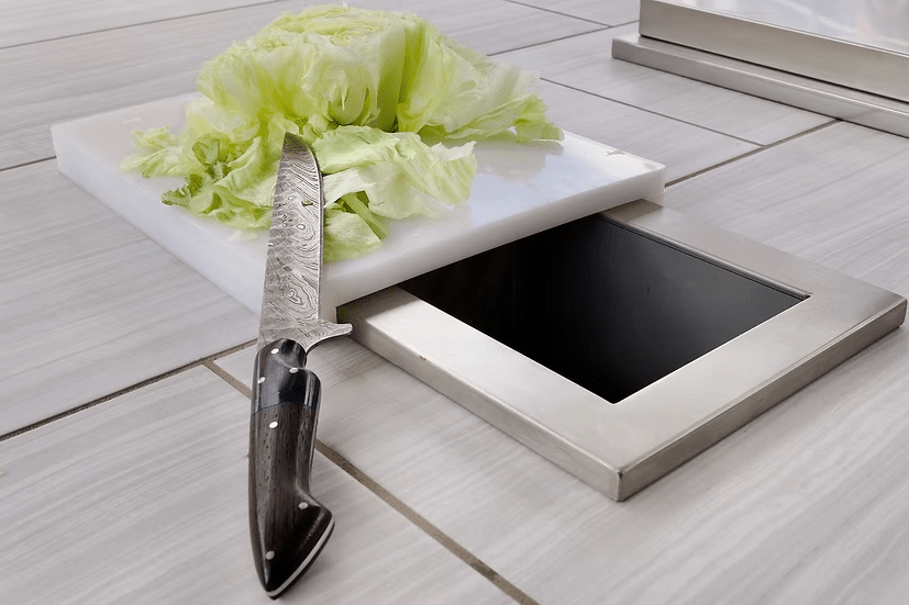 A head of lettuce and a knife sit on the Kokomo Grills Trash Chute and Cutting Board KO-TRSCH, placed on the tiled counter in an outdoor kitchen.