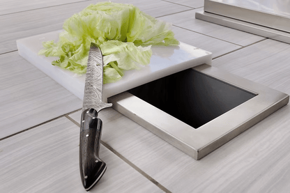 A head of lettuce and a knife sit on the Kokomo Grills Trash Chute and Cutting Board KO-TRSCH, placed on the tiled counter in an outdoor kitchen.