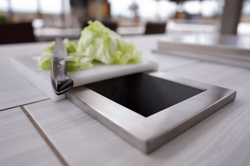 A knife and chopped lettuce sit on the Kokomo Grills Trash Chute and Cutting Board KO-TRSCH—a stainless steel trash chute embedded in a countertop—perfect for quick food scrap disposal in your outdoor kitchen setup.