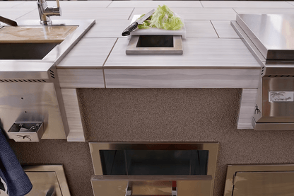 The Kokomo Grills Trash Chute and Cutting Board KO-TRSCH enhances this modern outdoor kitchen, featuring a built-in sink, cutting board, and trash chute. A knife lies next to chopped lettuce on a white tray above stainless steel drawers.