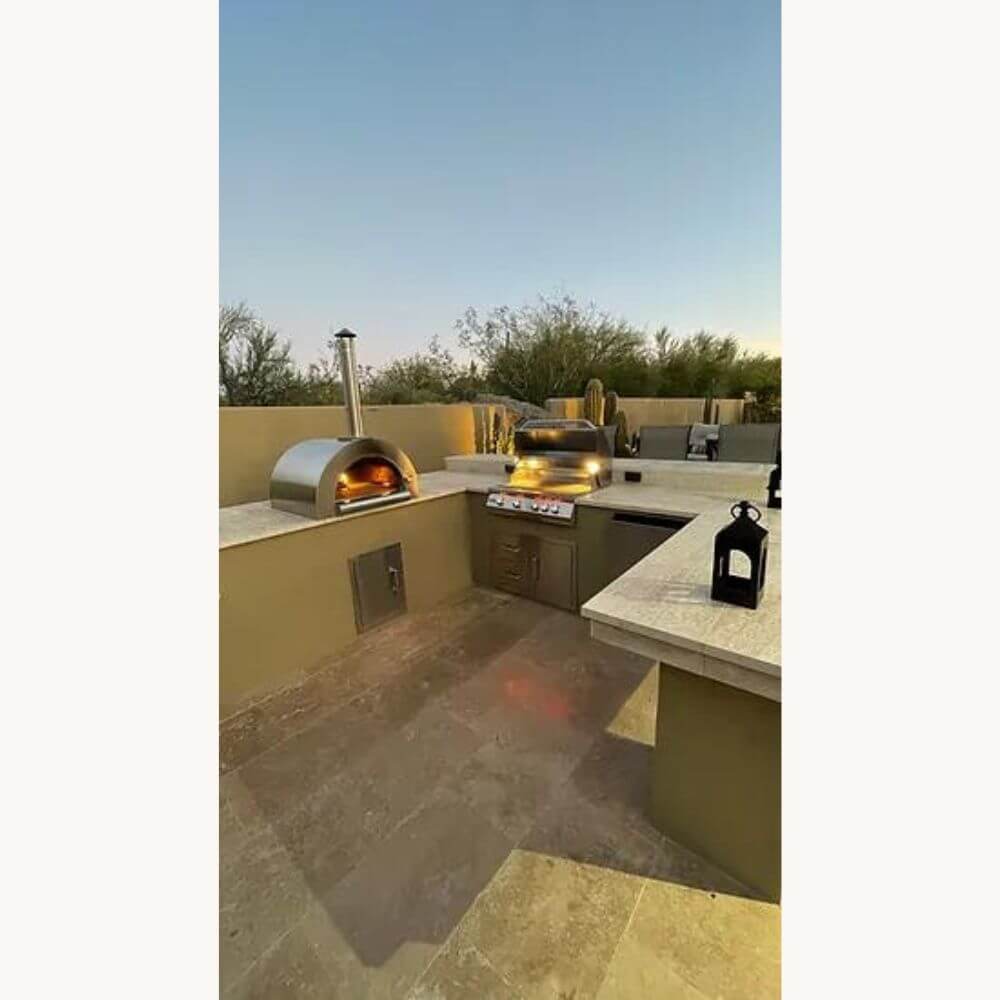 Actively burning Kokomo 32” Outdoor Wood-Fired Stainless Steel Pizza Oven on a marble countertop in a luxury outdoor kitchen, alongside a gas grill.