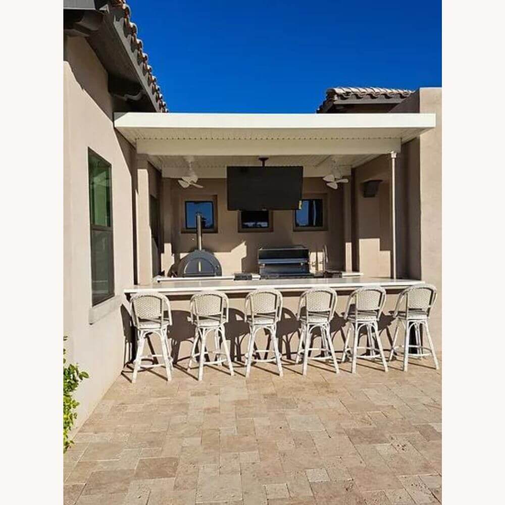 Outdoor kitchen with a bar counter and six stools, featuring a grill, the Kokomo 32” Outdoor Wood-Fired Stainless Steel Pizza Oven, and a mounted TV under a pergola.