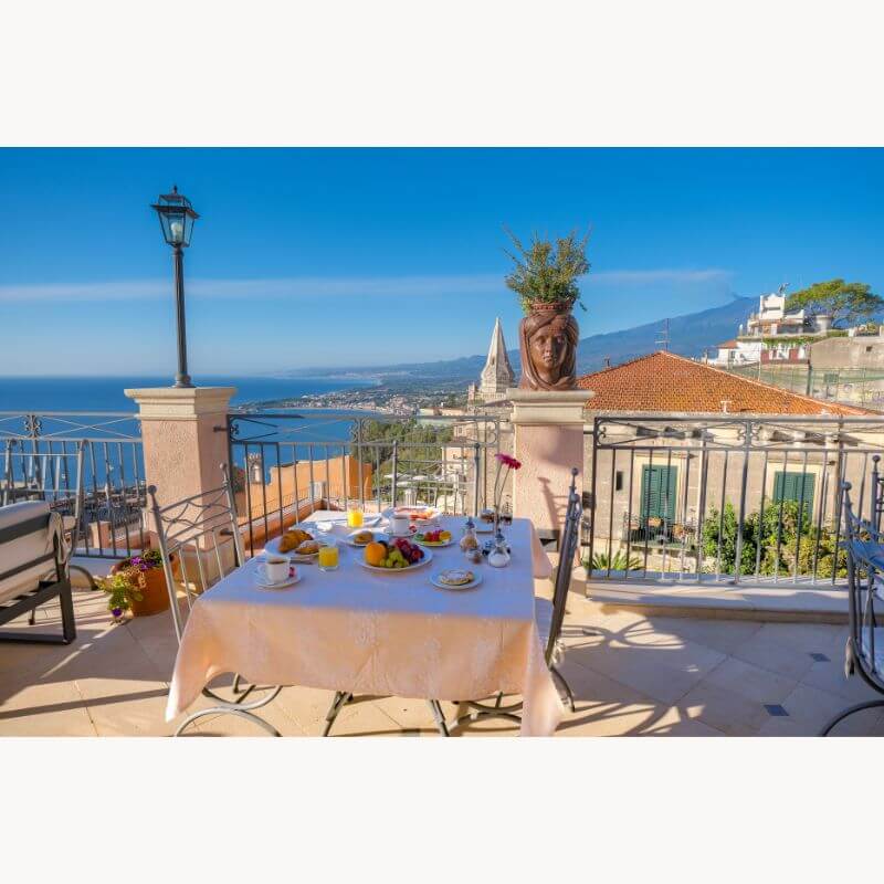 Outdoor dining table set for breakfast overlooking a coastal mediterranean town with sea and mountain views; clear blue sky in the background.