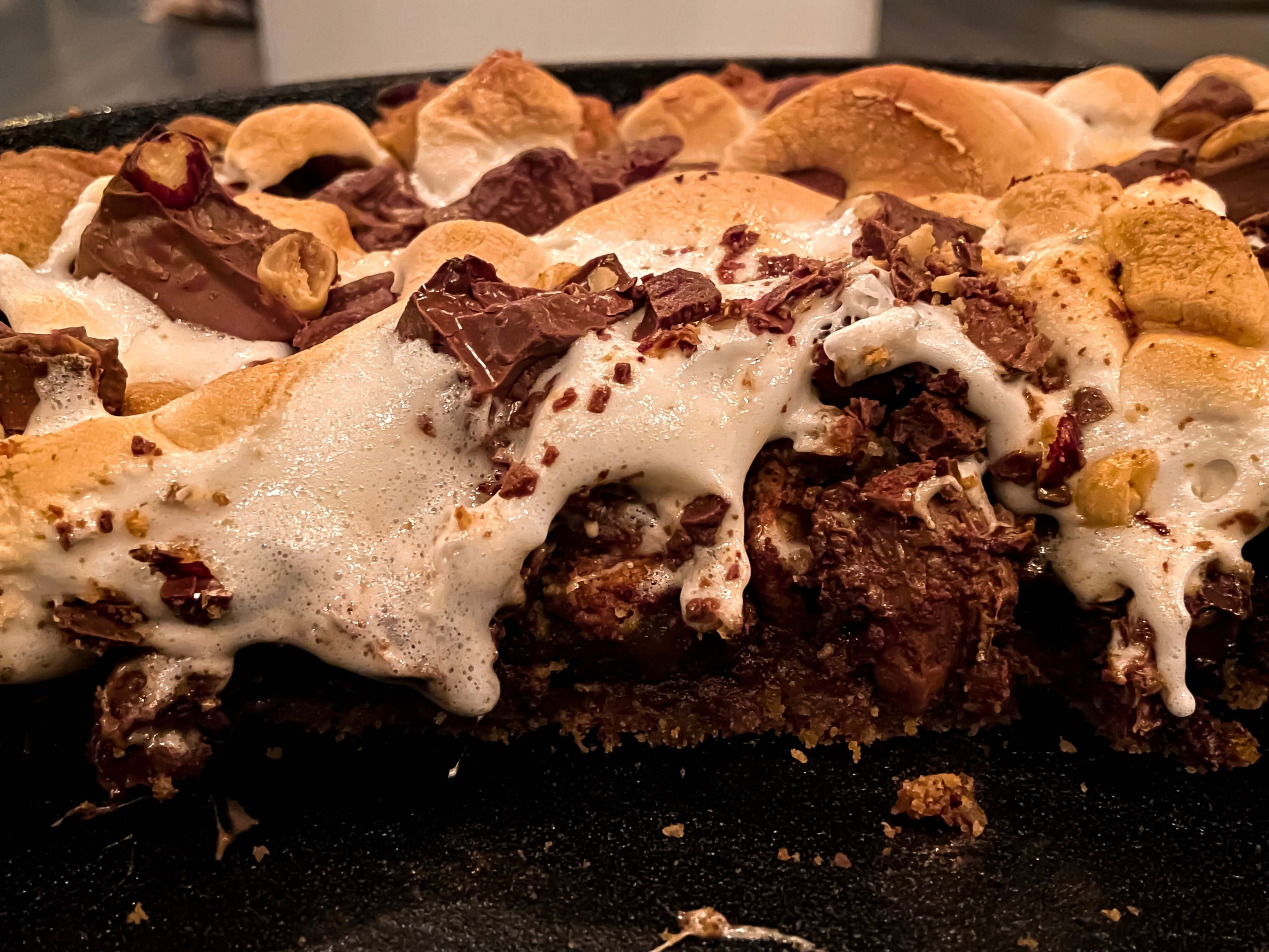 A wood-fired s'mores pie dessert with a graham cracker crust, melted chocolate, and golden-brown marshmallows. The chocolate and marshmallows are partially melted and dripping.