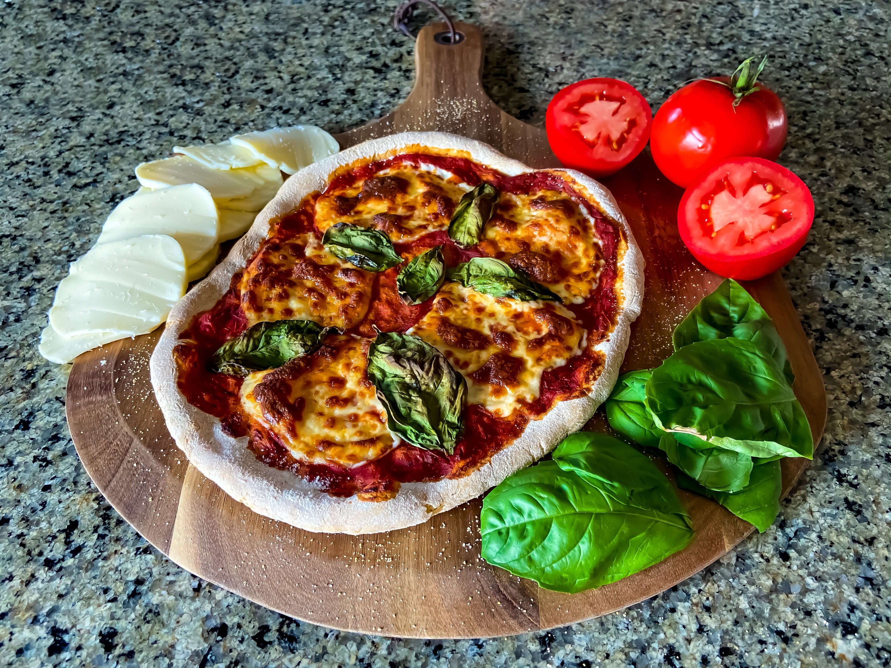 A margherita pizza on a wooden peel, surrounded by whole tomatoes, a stack of fresh mozzarella slices, and bay leaves on a marble countertop.