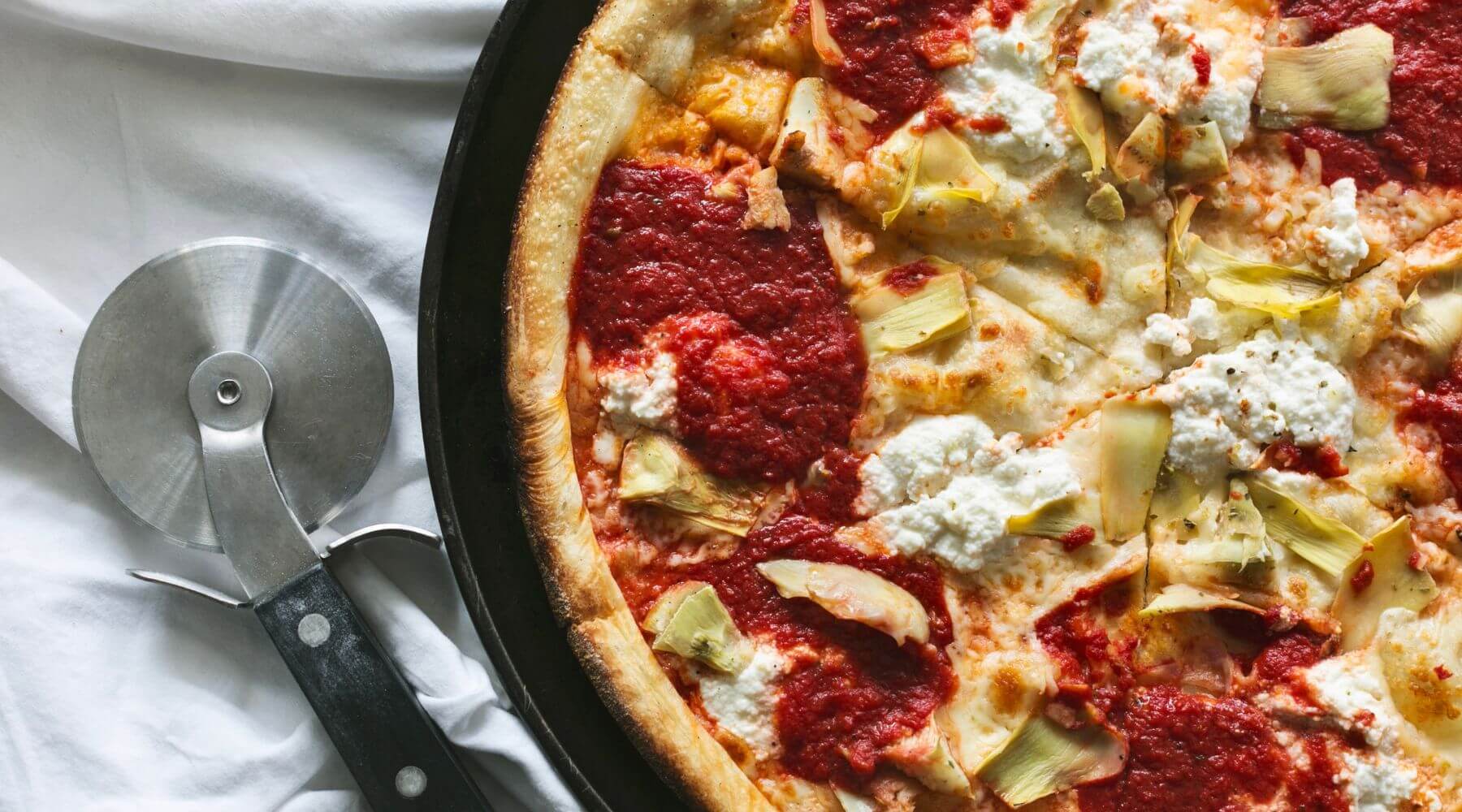 Pizza with artichokes, dollops of ricotta, and tomato sauce on a black pan, placed on a white cloth, alongside a round pizza cutter.