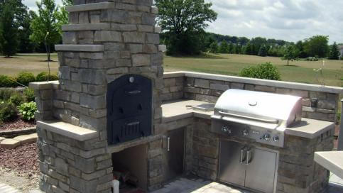 Tuscan Chef Medium Built-In Deluxe Family Pizza Oven (Model GX-CM) shown installed in a tall white stone enclosure with a firewood storage compartment beneath, on a patio outdoor kitchen.