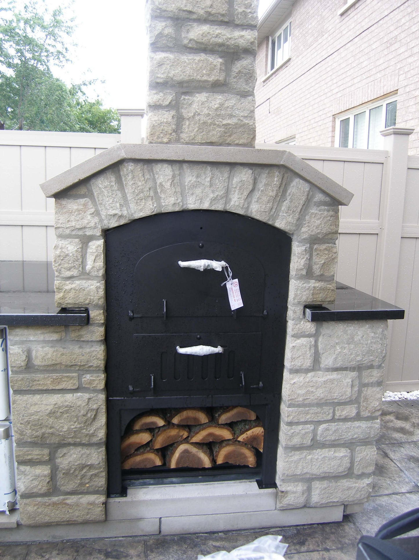 Tuscan Chef Medium Built-In Deluxe Family Pizza Oven (Model GX-CM), brand new with inspection tags still attached. The oven is housed in a white brick enclosure in a backyard. Fresh logs are neatly stacked in the lower unit's storage compartment.