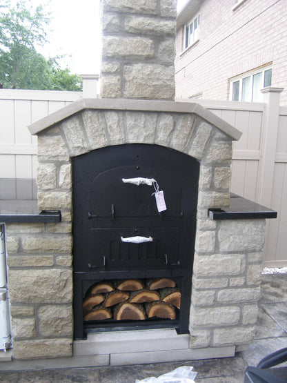 Tuscan Chef Medium Built-In Deluxe Family Pizza Oven (Model GX-CM), brand new with inspection tags still attached. The oven is housed in a white brick enclosure in a backyard. Fresh logs are neatly stacked in the lower unit's storage compartment.