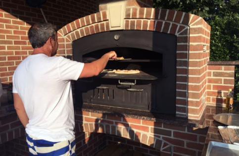 Amateur chef checking a pizza in his GX-DL Tuscan Chef oven in an outdoor kitchen on a sunny afternoon.