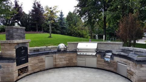 Outdoor stone kitchen with built-in GX-DL Tuscan Chef pizza oven, grill, sink, cabinets, and green lawn with trees in the background.