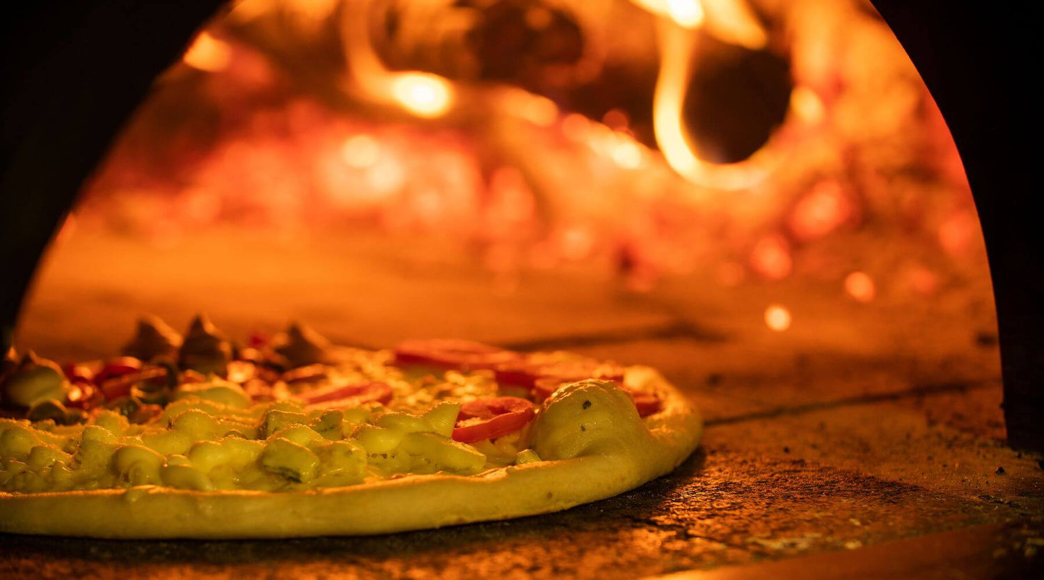 Pizza baking inside a wood-fired oven with visible flames and glowing embers in the background.