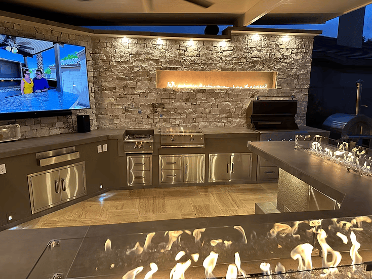 At night, the Kokomo Grills Luxury Outdoor Kitchen BBQ Grill Island shines with stainless steel appliances, a stone wall featuring a built-in linear fire, TV showing people in a pool, glass firepit on the counter, and warm lighting for a cozy setting.