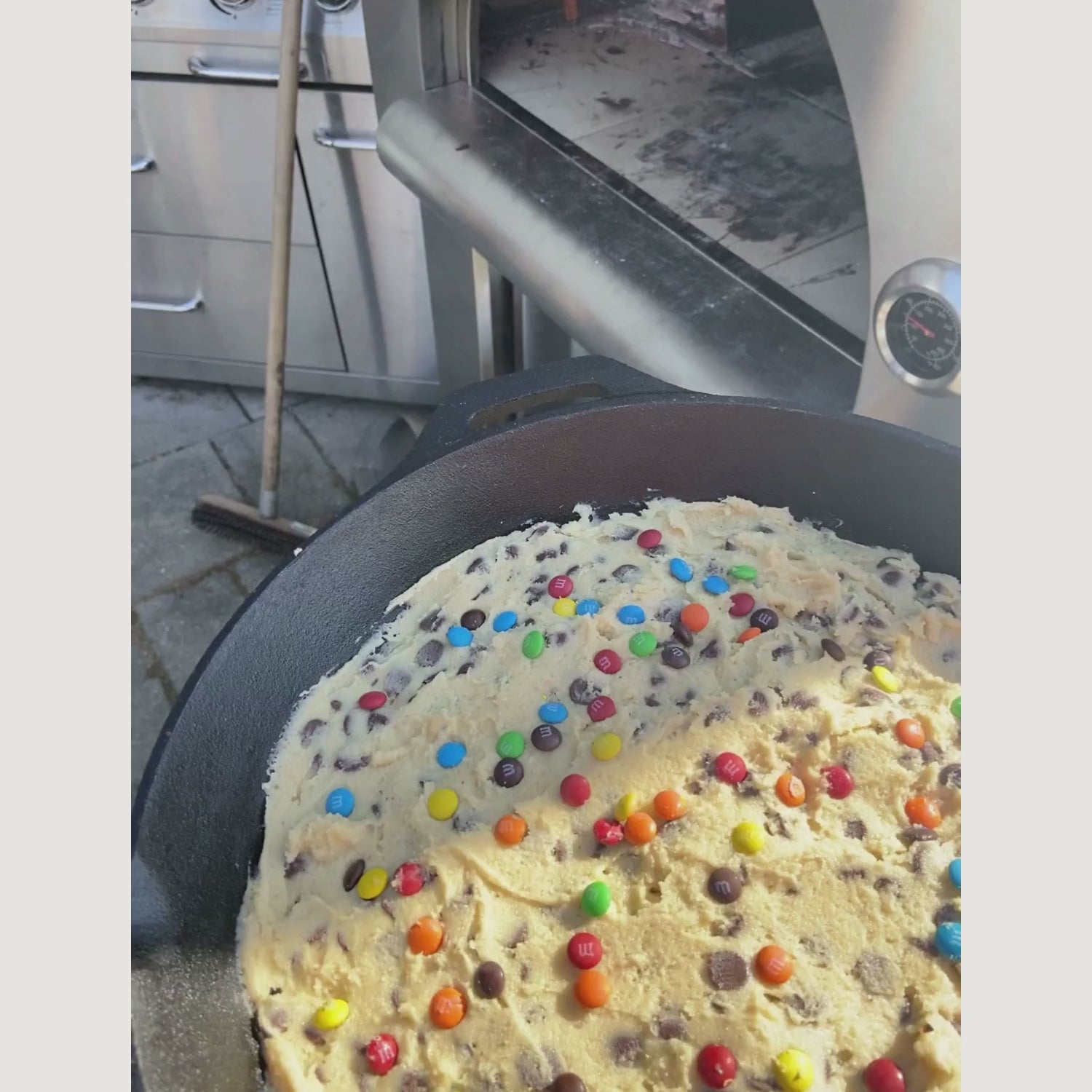 A person is inserting a raw cookie studded with colorful candies into a cast-iron skillet inside the burning PINNACOLO PREMIO Wood-Fired Outdoor Pizza Oven.