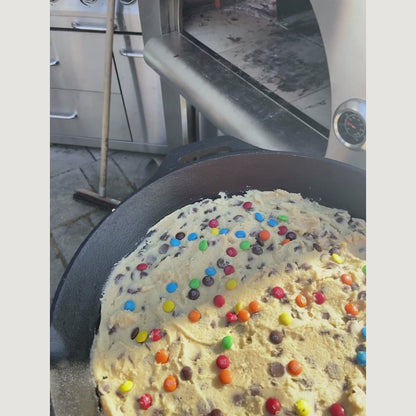 A person is inserting a raw cookie studded with colorful candies into a cast-iron skillet inside the burning PINNACOLO PREMIO Wood-Fired Outdoor Pizza Oven.
