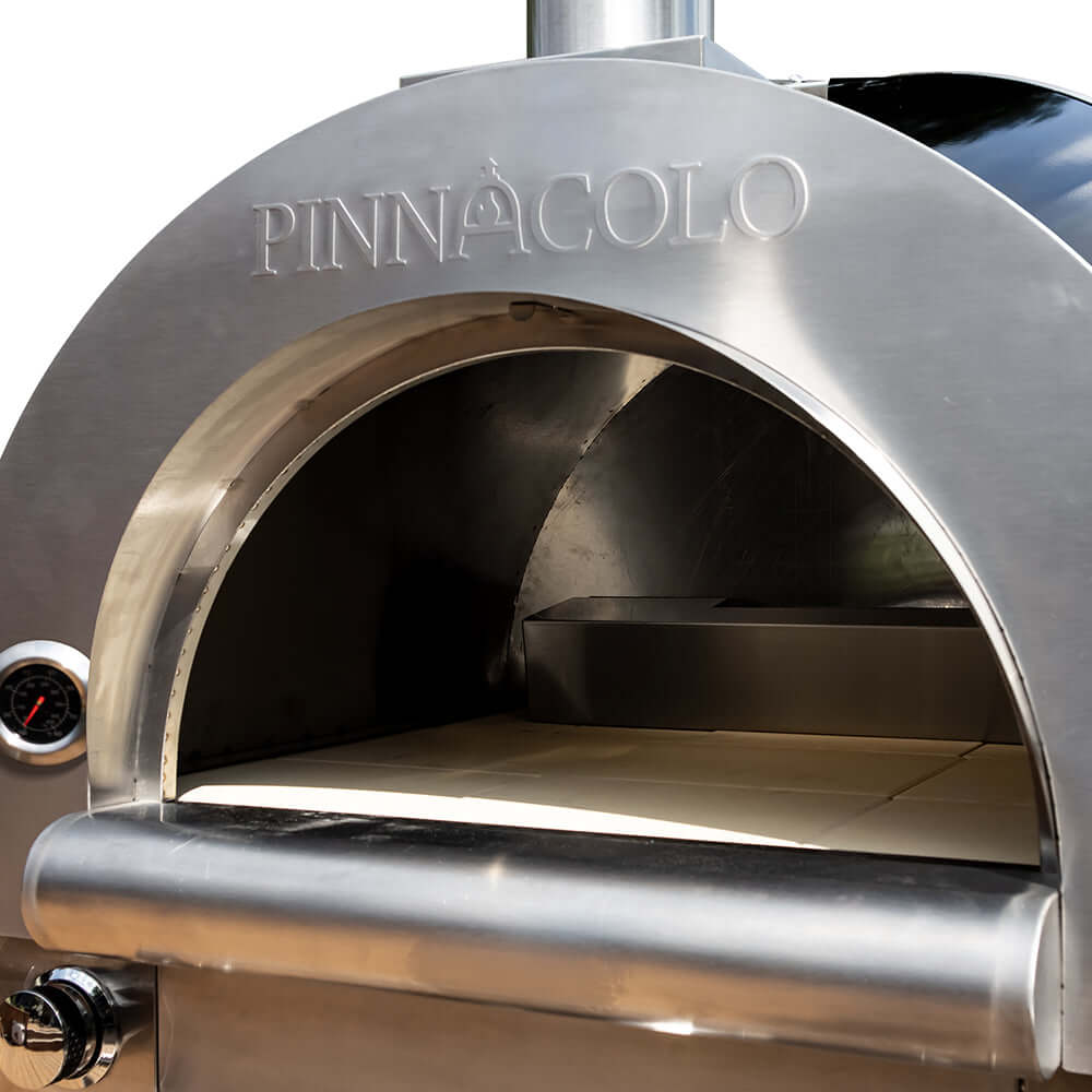 Close-up of the cooking chamber of the stainless steel PINNACOLO Fire One Up dual fuel pizza oven, showing the logo on top and white fire bricks below it.