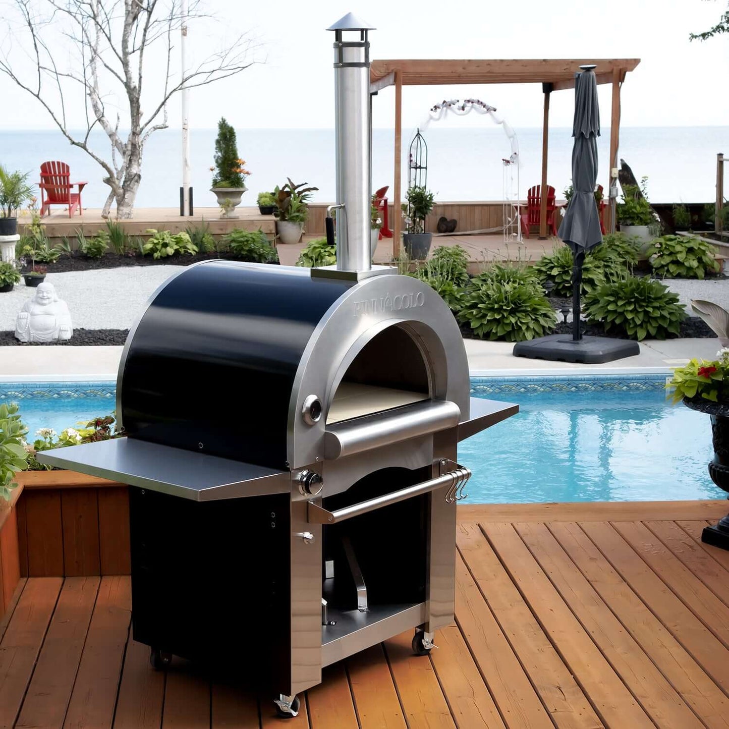 On a wooden deck by the pool, the Fire One Up PINNACOLO IBRIDO Wood/Gas Outdoor Pizza Oven stands ready on wheels. Deck chairs and a pergola frame a peaceful sea view, with lush plants beneath a bright sky.