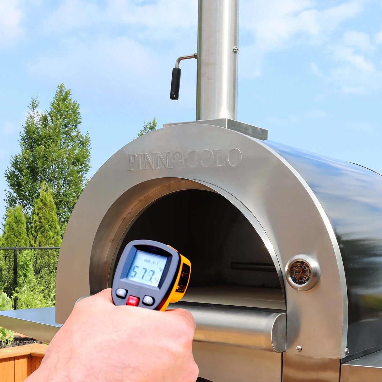 A hand holds the Fire One Up PINNACOLO IBRIDO Wood/Gas Outdoor Pizza Ovens infrared thermometer, displaying 577 degrees. Trees and a blue sky frame the scene.