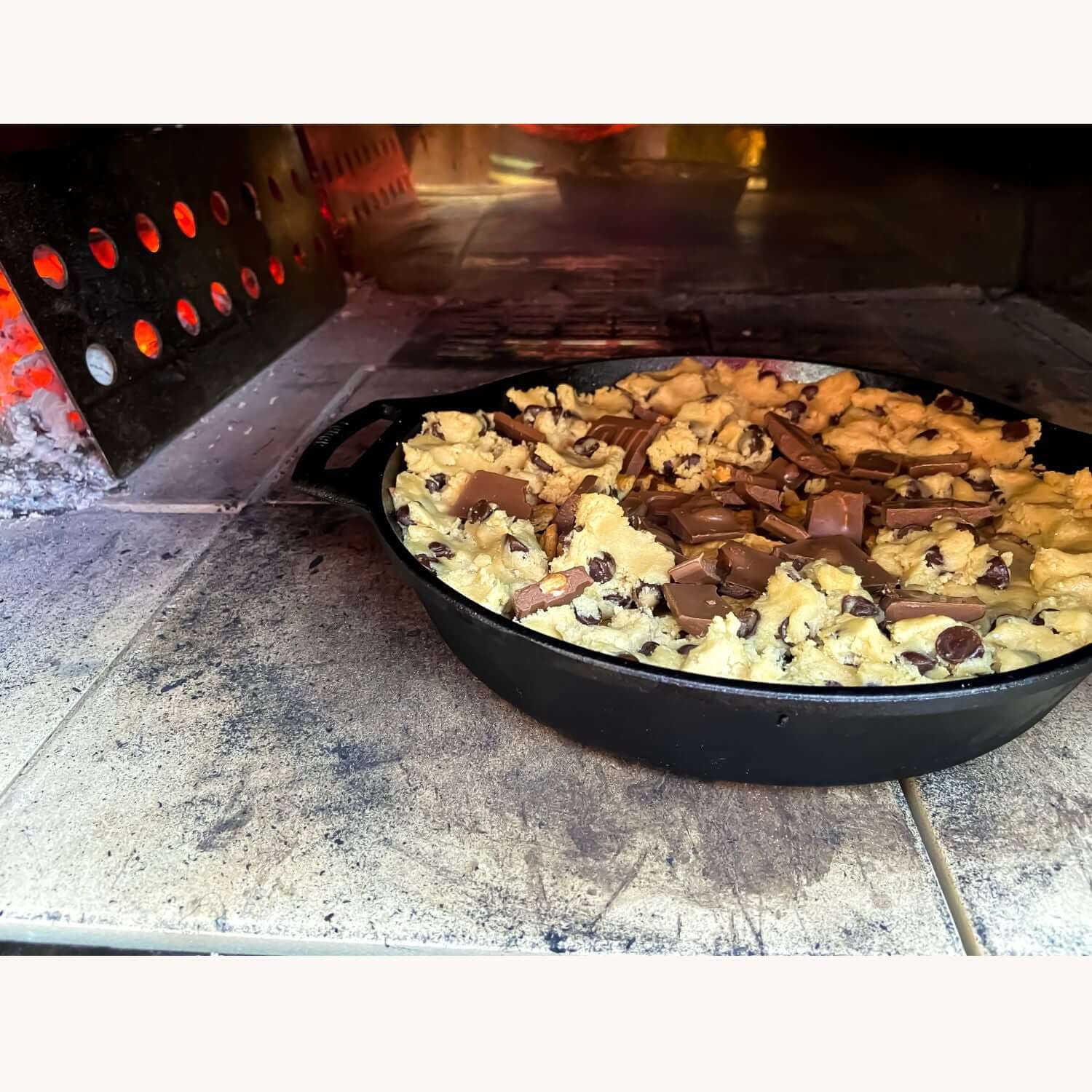 A PINNACOLO IBRIDO by Fire One Up bakes a S'mores pie in a cast-iron skillet with chocolate chunks. The hybrid wood/gas pizza oven's visible flame flickers on the left.