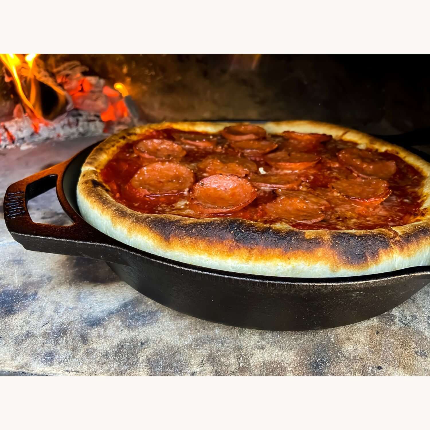 A pepperoni pizza with a thick crust sizzles in a cast iron pan inside the Fire One Up PINNACOLO IBRIDO (Hybrid) Wood/Gas Outdoor Pizza Oven. The edges are slightly charred, as vibrant flames flicker in the background.