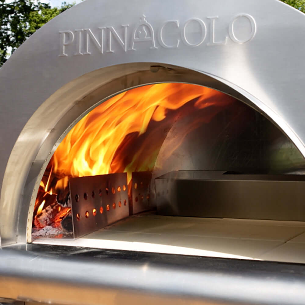 The Fire One Up PINNACOLO IBRIDO stainless steel outdoor pizza oven with roaring wood-fire flames heating its cordierite cooking surface; compatible with wood or gas fuel.