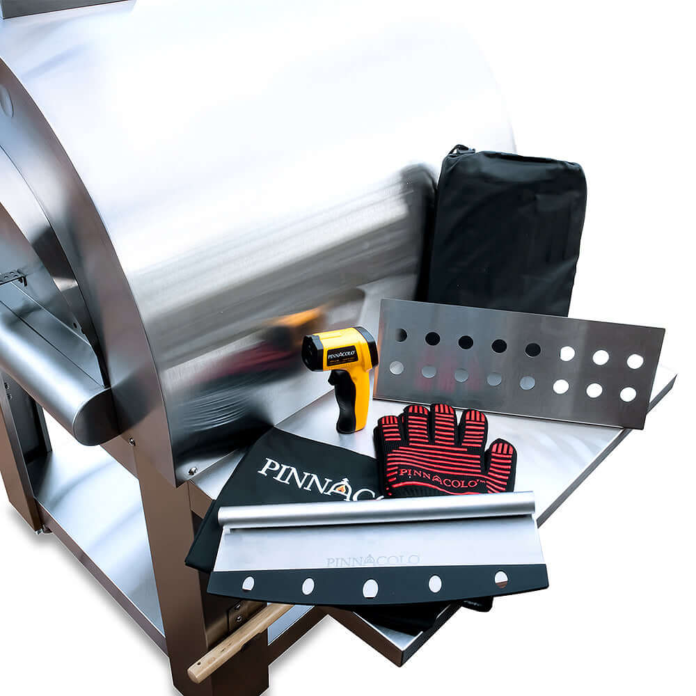 Overhead view of the PINNACOLO PREMIO Wood-Fired Outdoor Pizza Oven with accessories on its side shelf, including an infrared thermometer, protective gloves, fire separator, cutter, and cover.