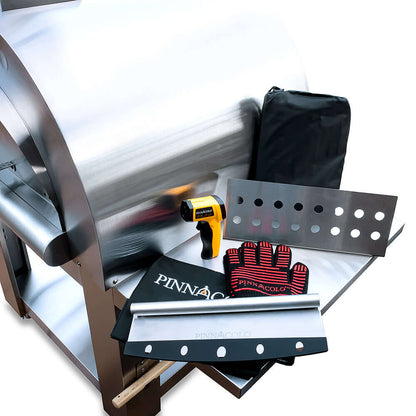 Overhead view of the PINNACOLO PREMIO Wood-Fired Outdoor Pizza Oven with accessories on its side shelf, including an infrared thermometer, protective gloves, fire separator, cutter, and cover.