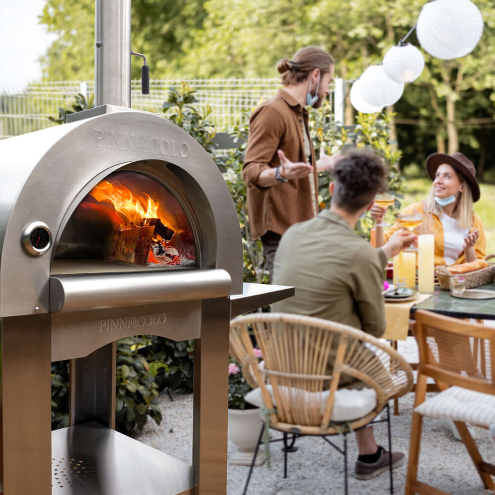 Three people enjoy an outdoor gathering near the PINNACOLO PREMIO Wood-Fired Outdoor Pizza Oven, surrounded by trees and string lights. One person stands, gesturing, while the others sit at a table with food and drinks, engaged in conversation.