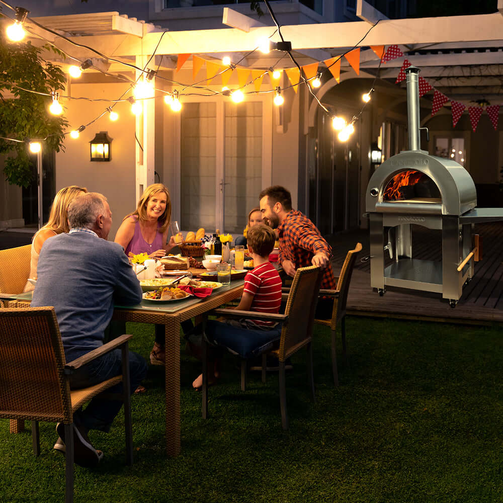 A family sits around a table in a backyard at night, smiling and engaged in conversation, enjoying a meal under string lights. The PINNACOLO PREMIO Wood-Fired Outdoor Pizza Oven is burning nearby.