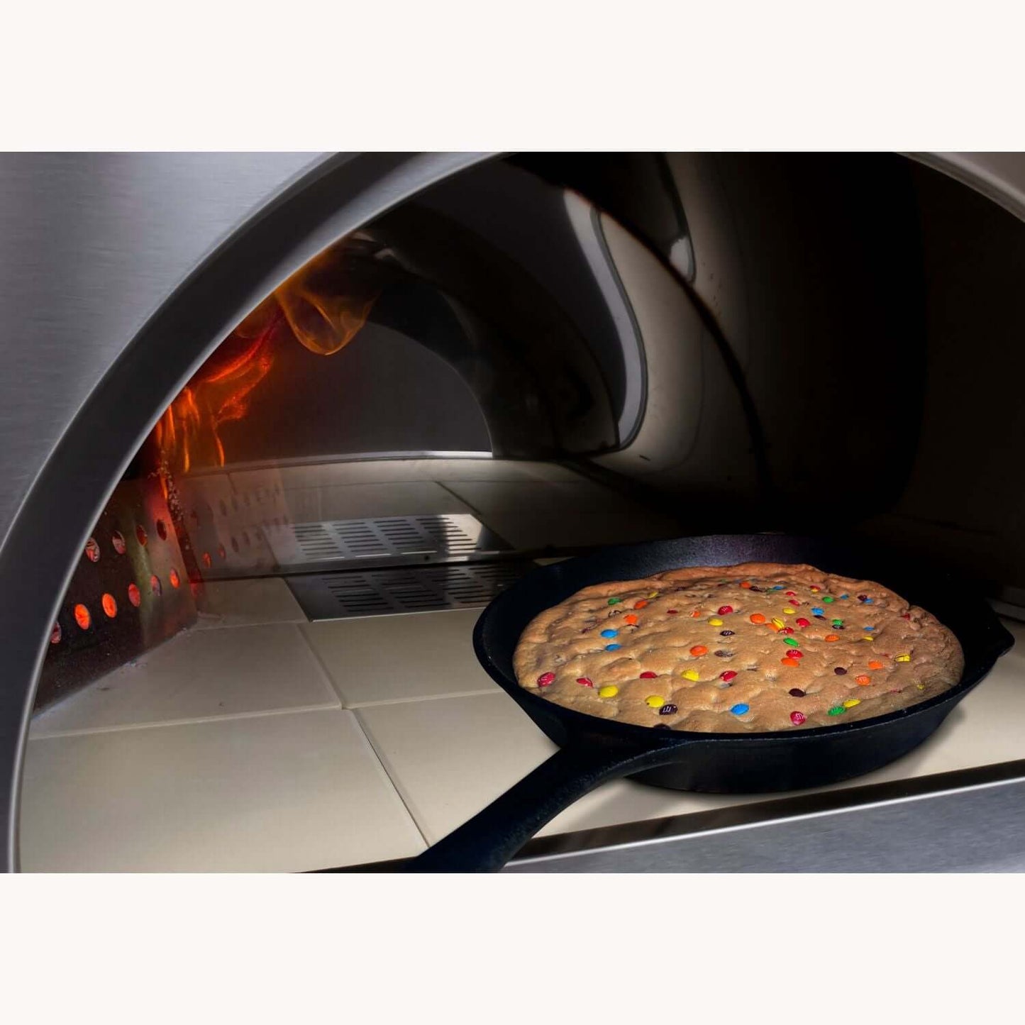 A large cookie studded with colorful candies is baking in a cast-iron skillet inside the PINNACOLO PREMIO Wood-Fired Outdoor Pizza Oven, with flames shooting upward.