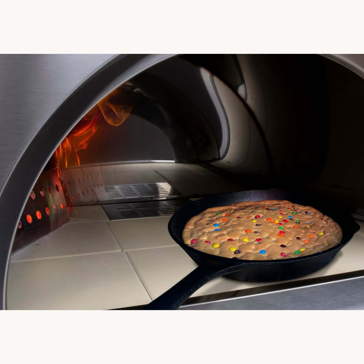 A large cookie studded with colorful candies is baking in a cast-iron skillet inside the PINNACOLO PREMIO Wood-Fired Outdoor Pizza Oven, with flames shooting upward.