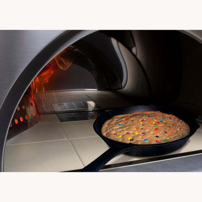 A large cookie studded with colorful candies is baking in a cast-iron skillet inside the PINNACOLO PREMIO Wood-Fired Outdoor Pizza Oven, with flames shooting upward.