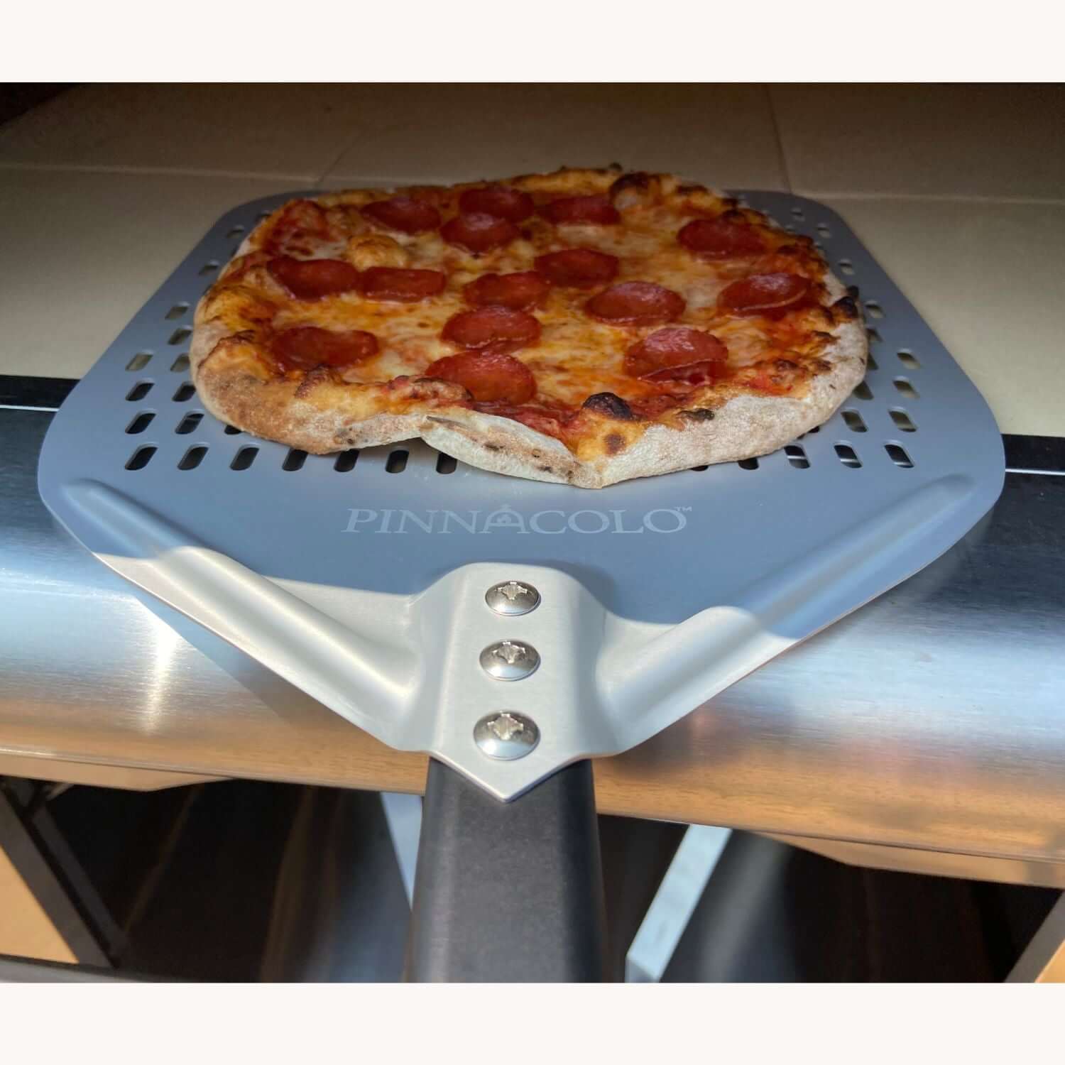 A hot pepperoni pizza is being pulled out of the PINNACOLO PREMIO Wood-Fired Outdoor Pizza Oven using a large, branded 'PINNACOLO' peel.