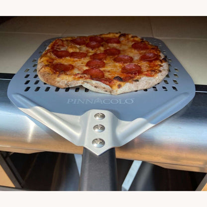 A hot pepperoni pizza is being pulled out of the PINNACOLO PREMIO Wood-Fired Outdoor Pizza Oven using a large, branded 'PINNACOLO' peel.