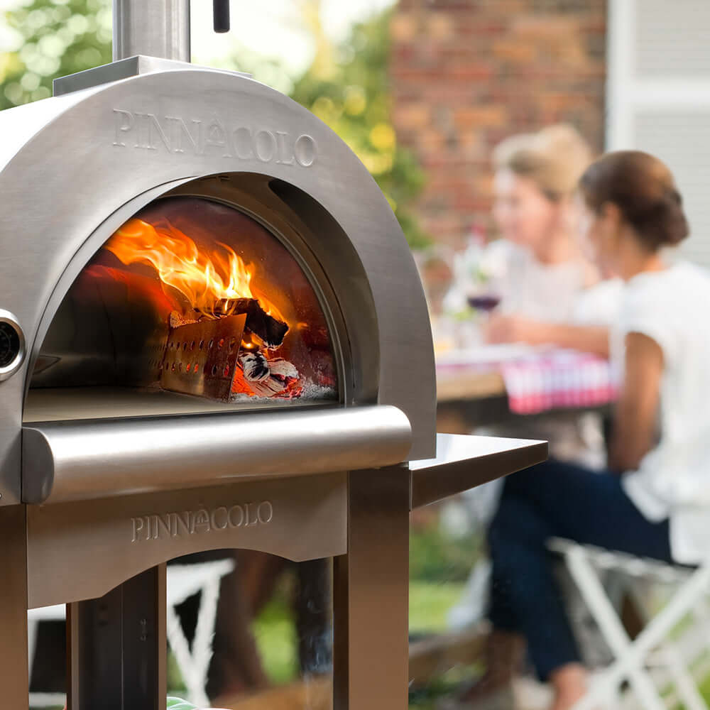 Close-up of the rolling flames inside the PINNACOLO PREMIO Wood-Fired Outdoor Pizza Oven, with people celebrating in the background.
