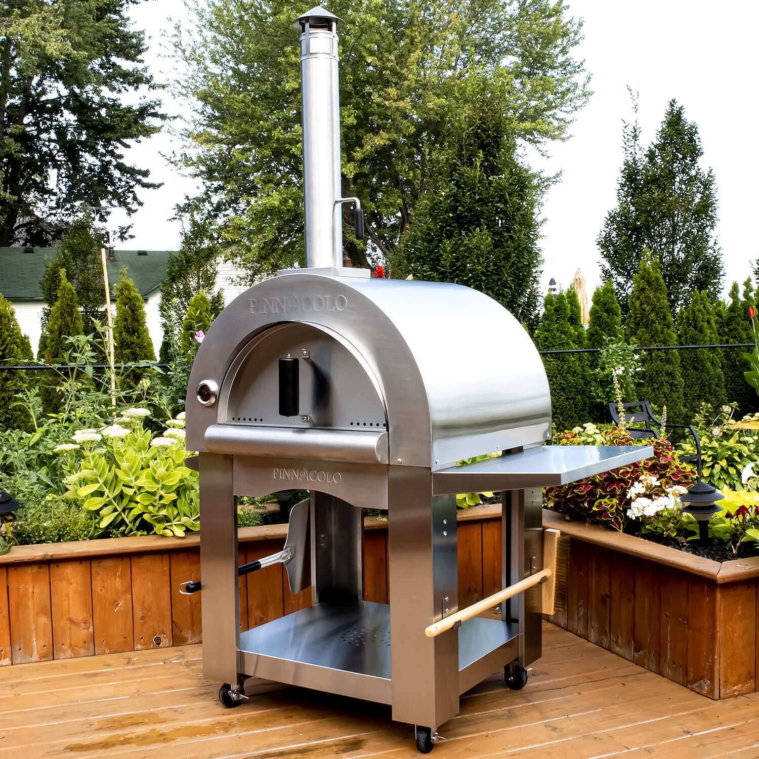 PINNACOLO PREMIO Wood-Fired Outdoor Pizza Oven, standing proudly on a wooden deck with a brush and paddle hanging beside it. Lush greenery and trees provide the perfect backdrop for alfresco dining.